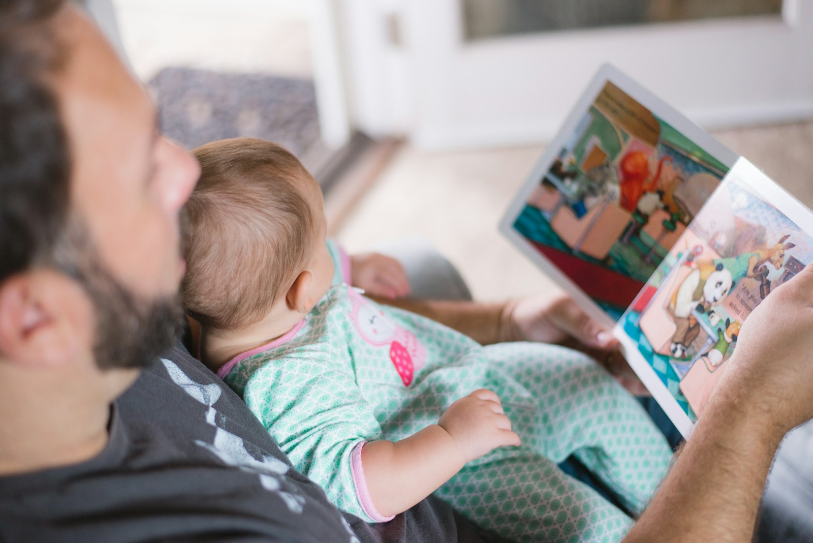 A father reads a picture book with his young child in the family living room.